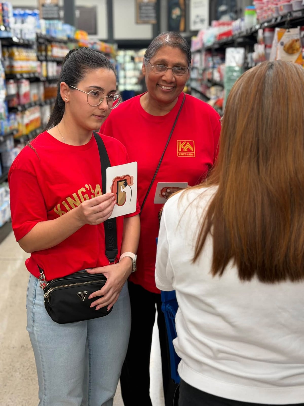 Two women in red King’s Army shirts share the Jesus At The Door card in a grocery-store conversation.