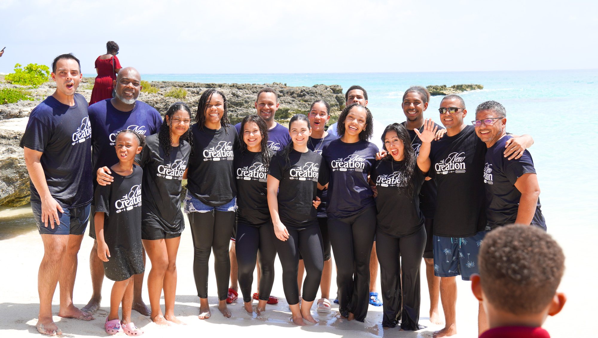 A group in New Creation shirts stands together on the beach after baptisms.