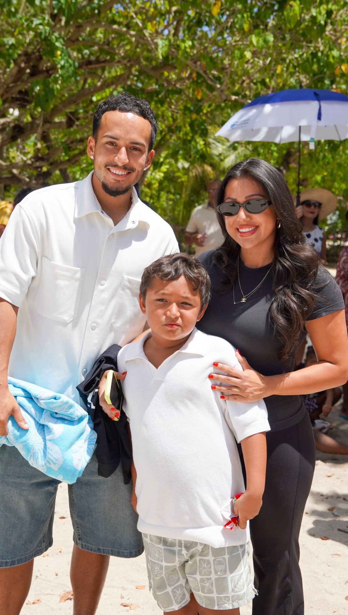 Two adults and a child stand together beneath trees beside the beach.