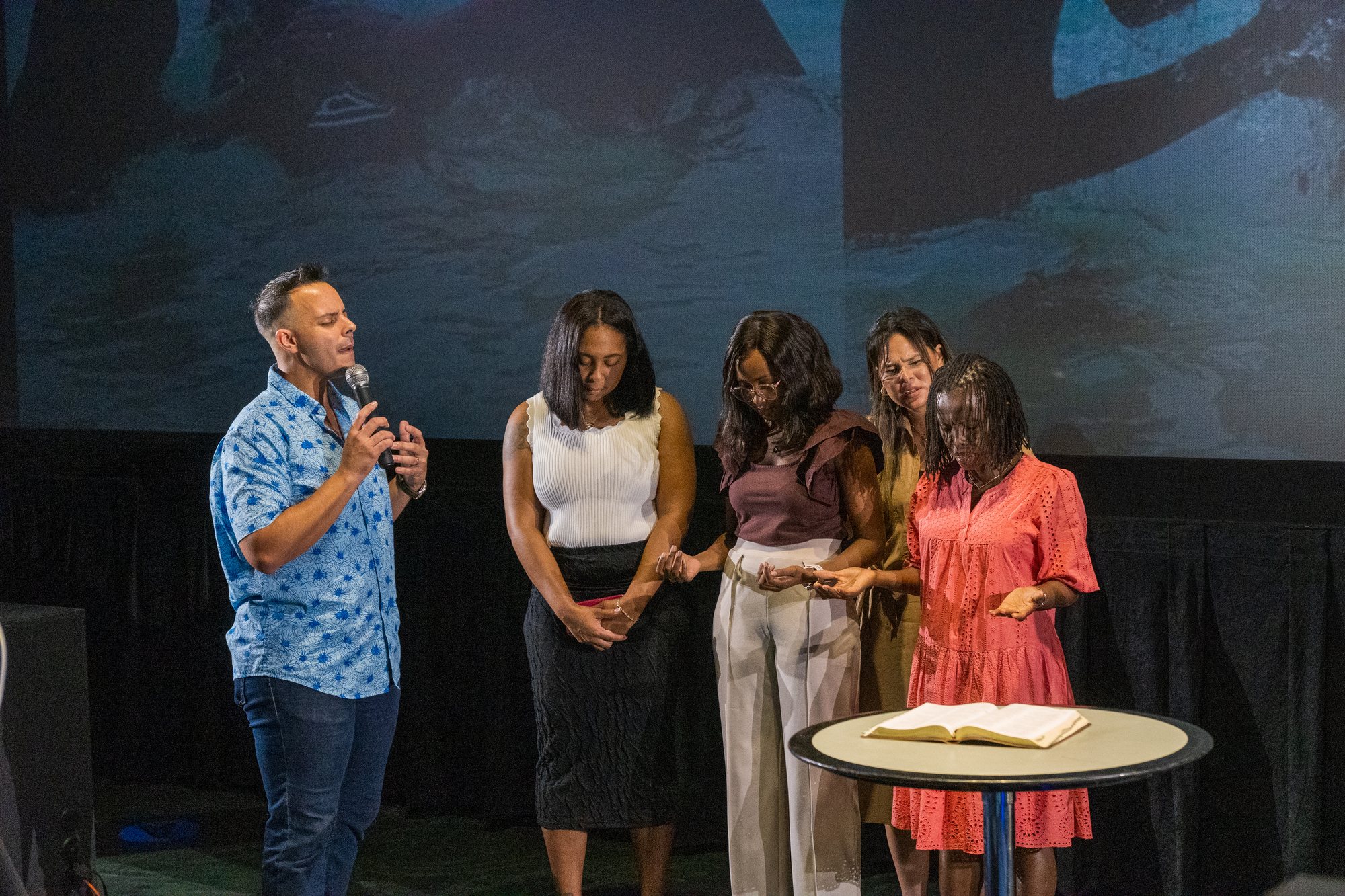 A speaker and four people stand with heads bowed beside an open Bible.