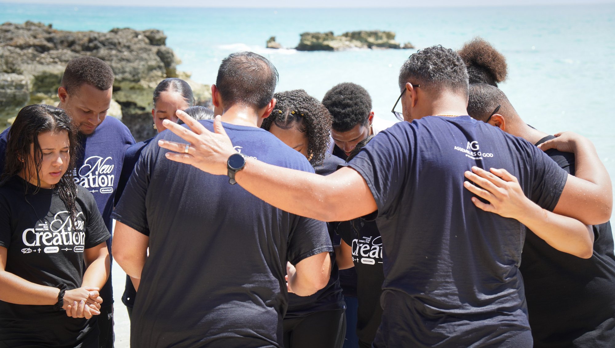 People stand close with arms around one another and heads bowed beside the sea.