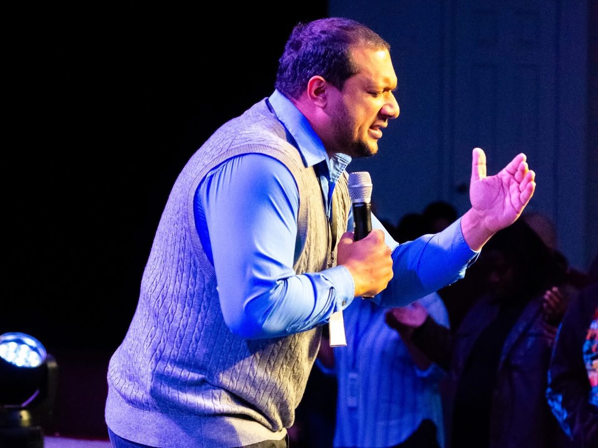 Pastor Jose Rivera kneeling in prayer on stage, microphone in hand